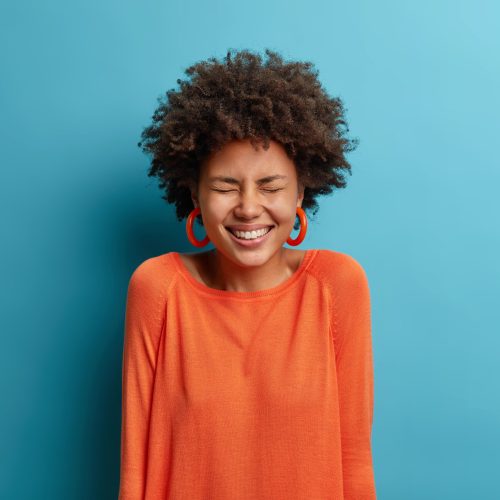 Upbeat cheerful African American woman laughs with joy, watches funny movie with friend, has carefree expression, talks casually in pub, dressed in orange jumper, isolated on blue background
